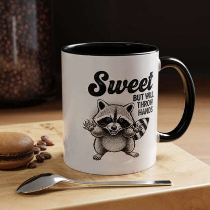 Mug with raccoon illustration and text on a wooden surface with coffee beans and a spoon.