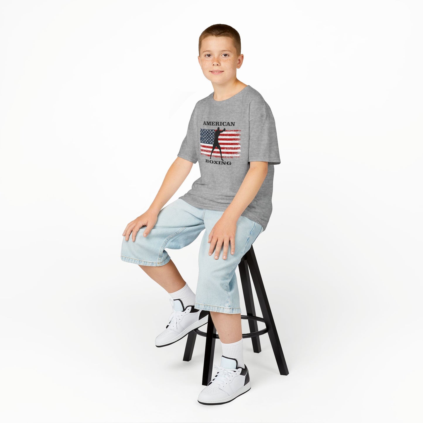 Young boy sitting on a stool wearing a gray t-shirt, light blue jeans, and white sneakers on a white background