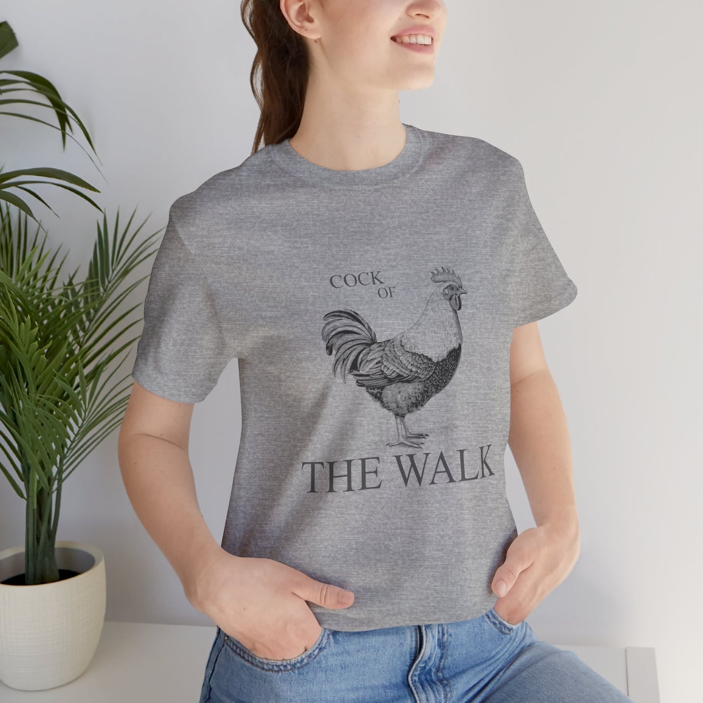 Person wearing a gray t-shirt with a rooster graphic and text, standing indoors next to a plant.
