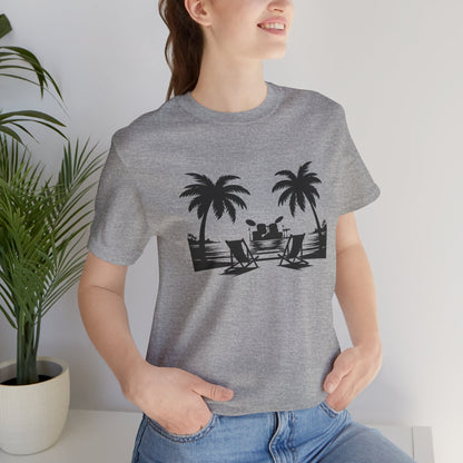 Person wearing a gray t-shirt with a black palm tree and beach chair design, standing indoors next to a plant.