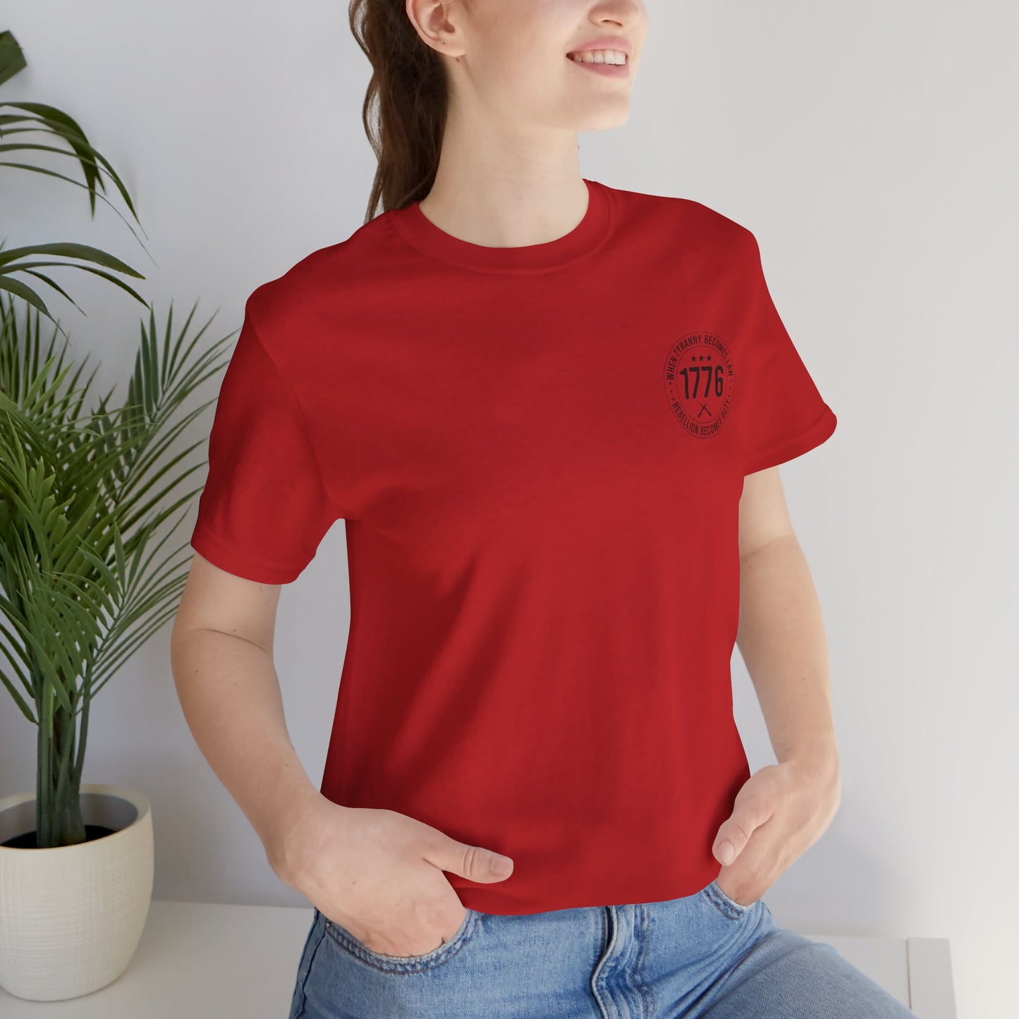 Person wearing a red t-shirt with a logo, standing indoors next to a plant.