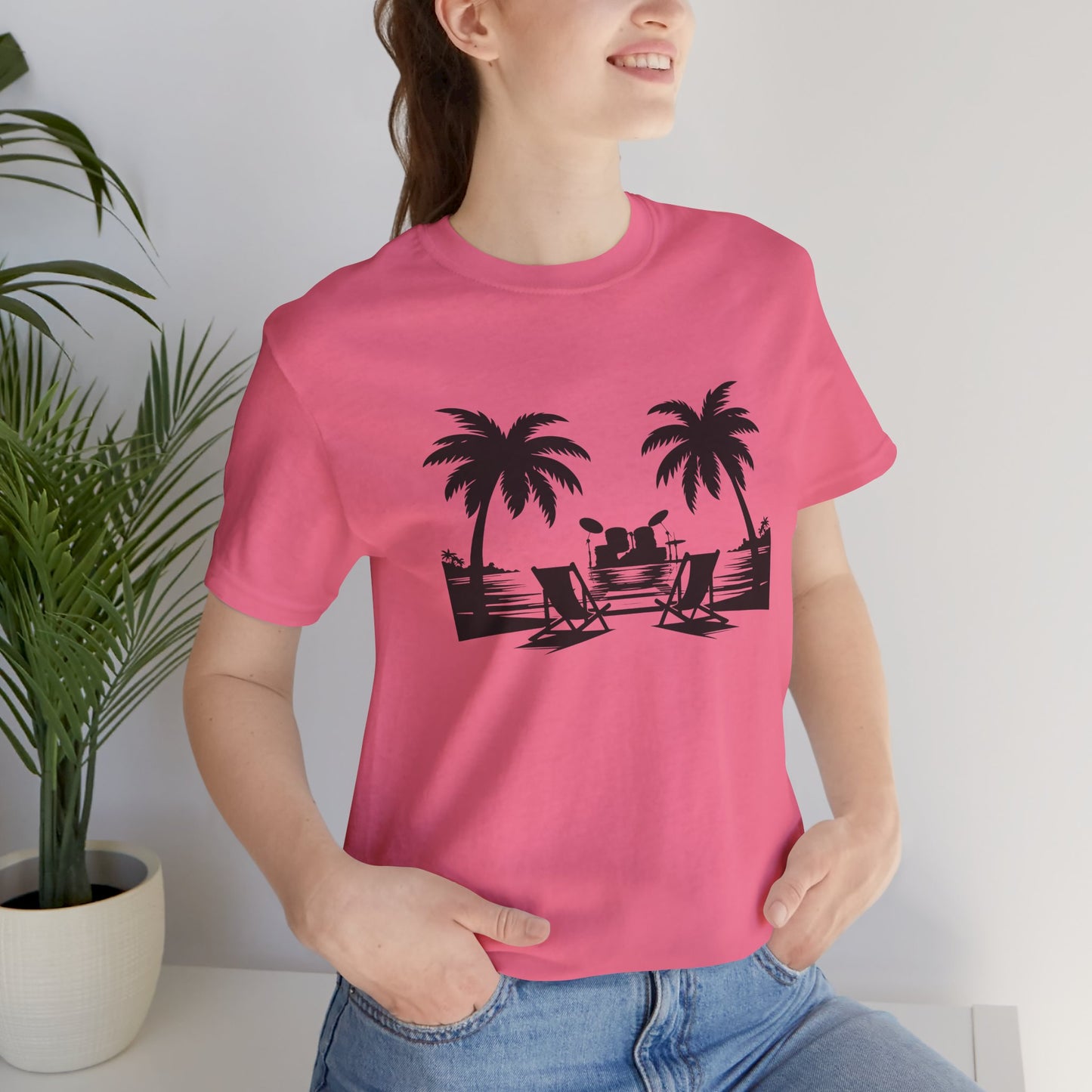 Person wearing a pink t-shirt with black palm tree and beach chair design, standing indoors next to a plant.