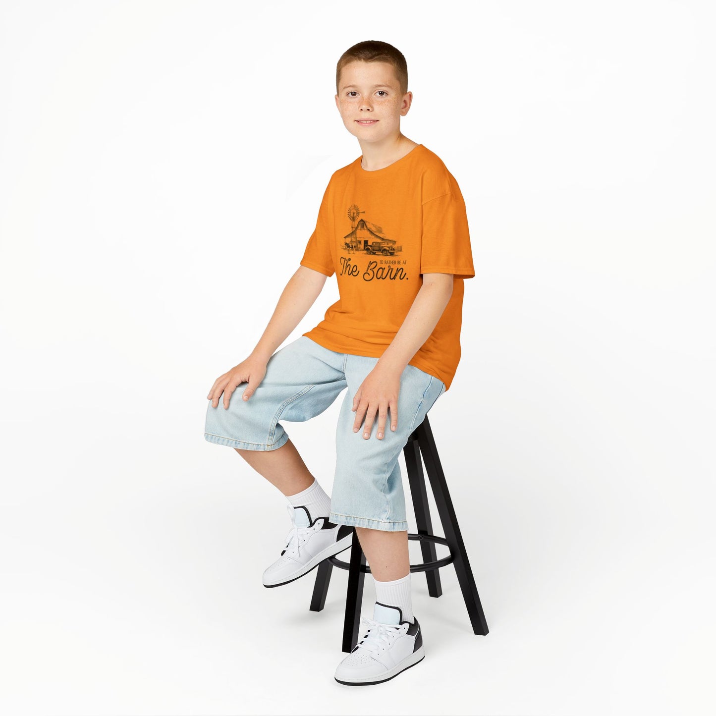 Child sitting on a stool wearing an orange t-shirt and light blue shorts on a white background