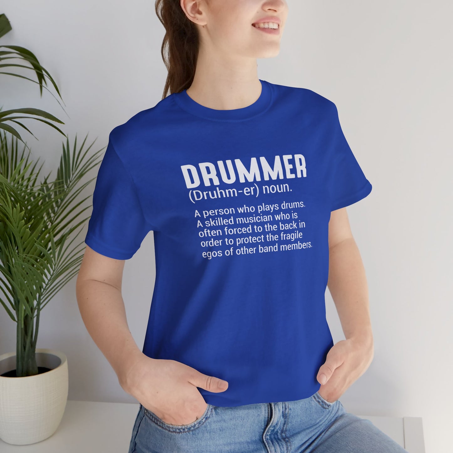 Person wearing a blue t-shirt with a definition of 'drummer' text, standing indoors with a plant in the background.