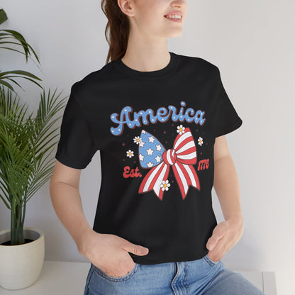 Person wearing a black t-shirt with an American-themed design, standing indoors next to a plant.