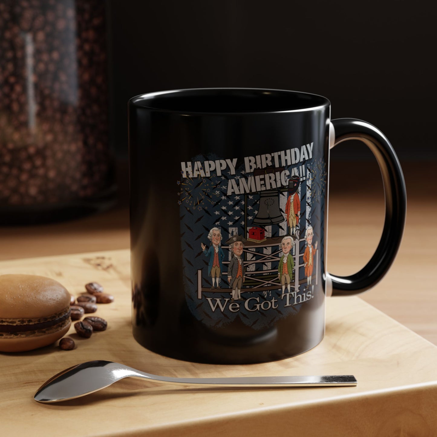 Black mug with 'Happy Birthday America! We Got This' design on a wooden surface with coffee beans and a macaron.
