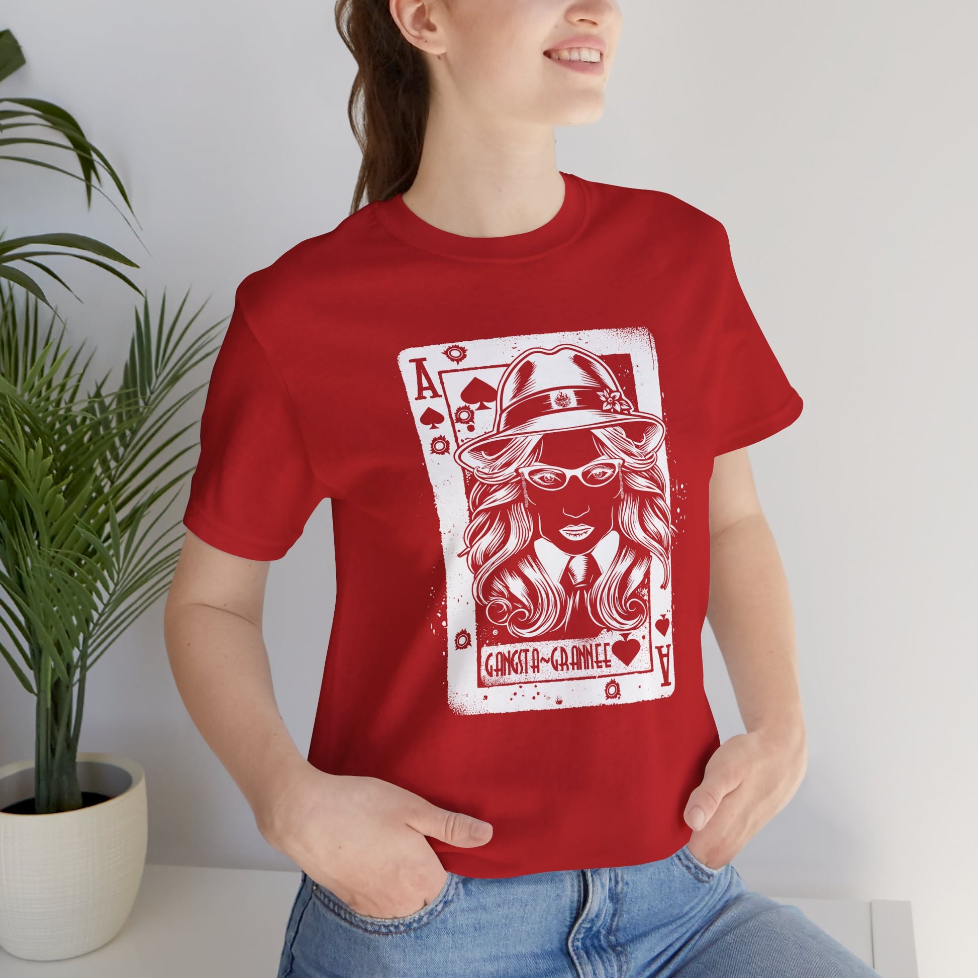 Person wearing a red t-shirt with a graphic design of a woman in a hat and glasses, sitting in a room with a plant.