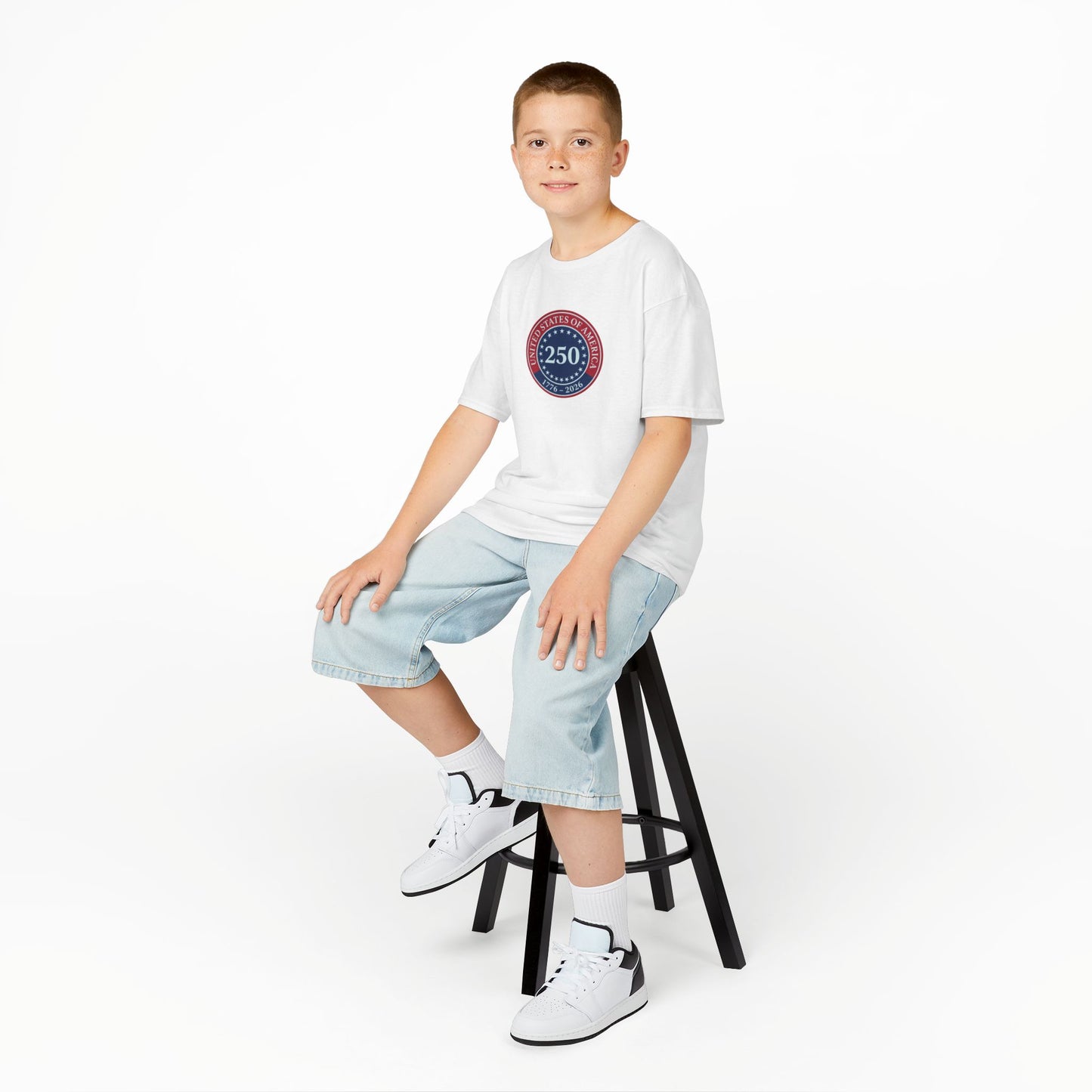 Young person sitting on a stool wearing a white t-shirt with text and graphics, light blue jeans, and white sneakers.