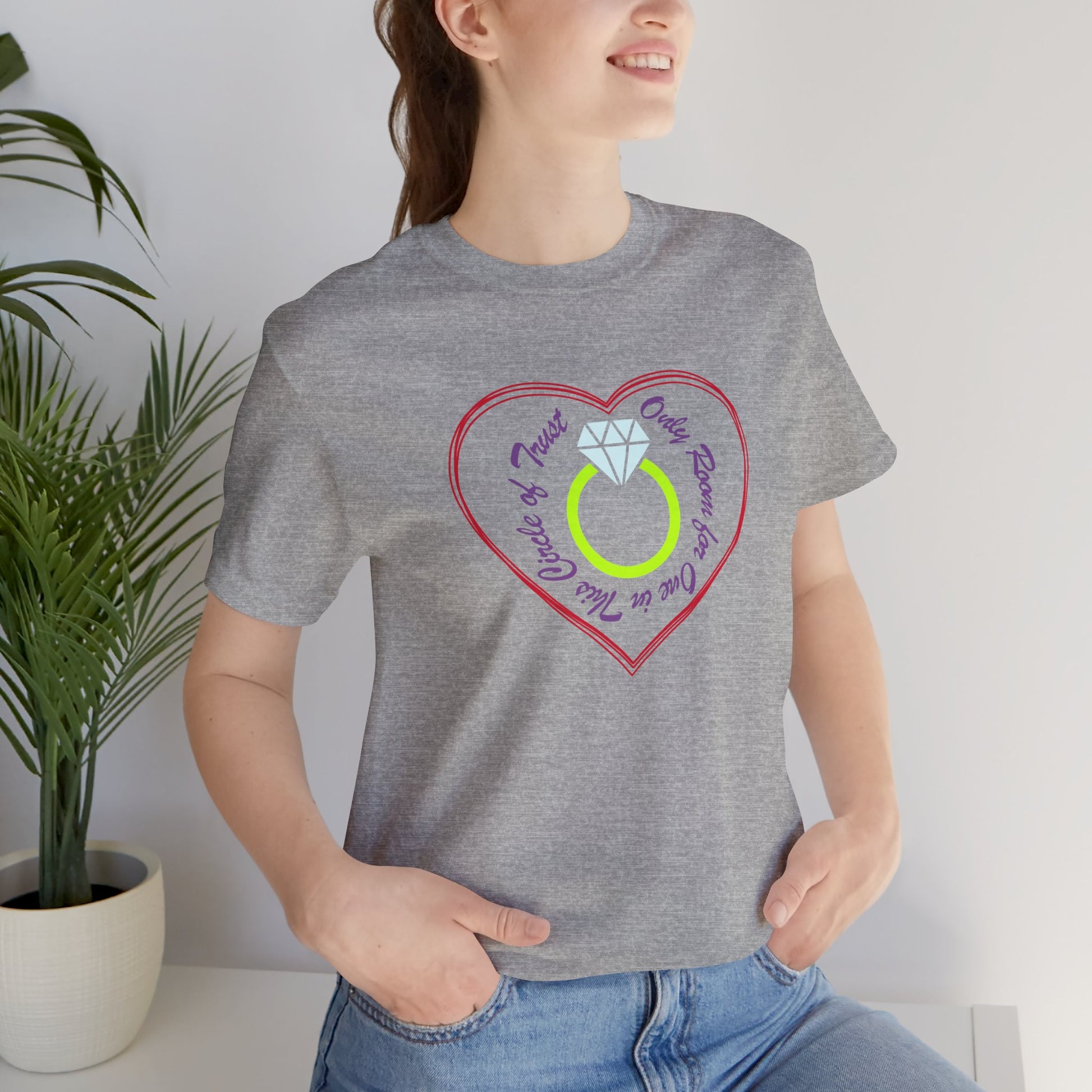 Person wearing a gray t-shirt with a heart-shaped design featuring a ring, standing indoors next to a plant.
