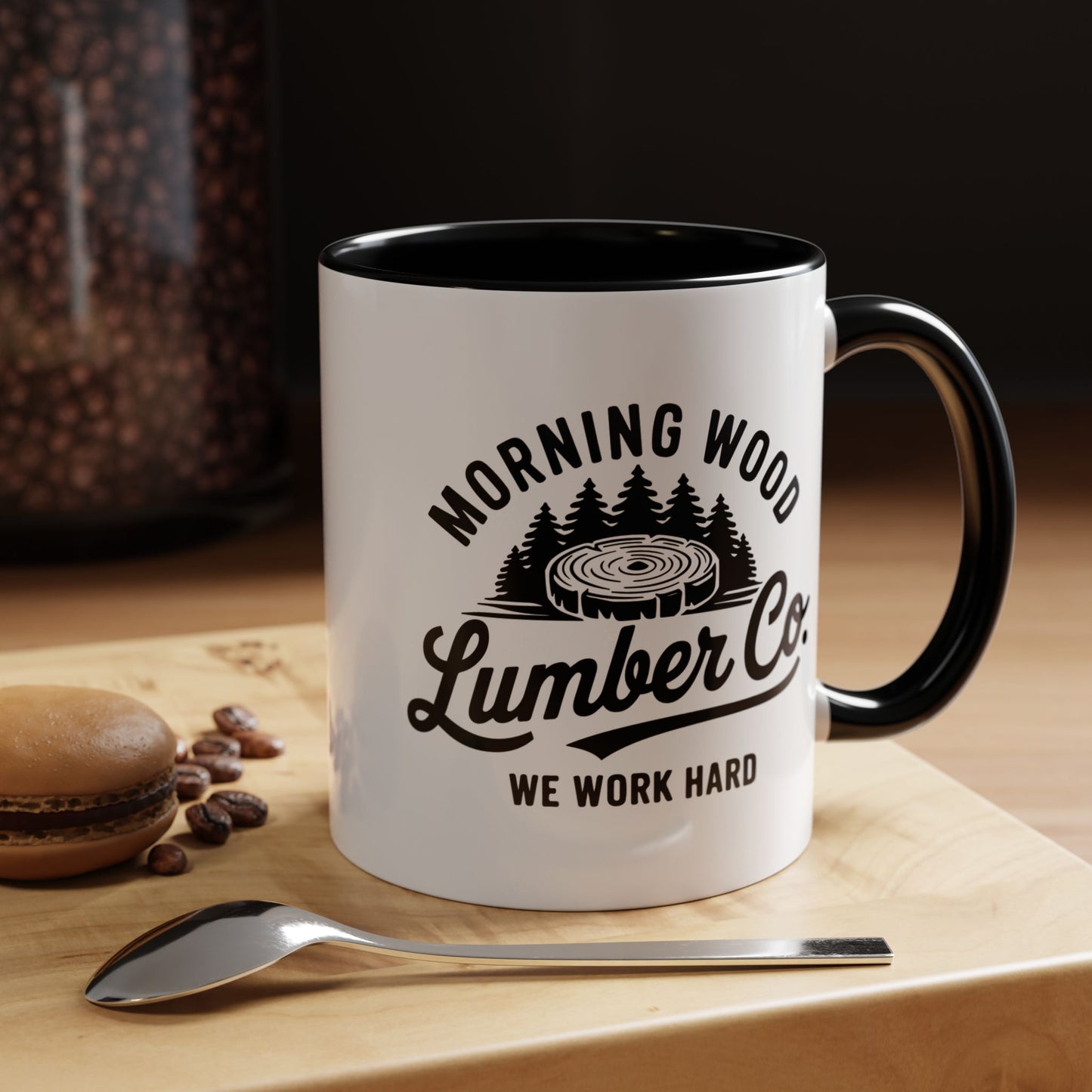 White mug with black interior and handle, featuring 'Morning Wood Lumber Co. We Work Hard' text and graphic, on a wooden surface with coffee beans and a spoon.