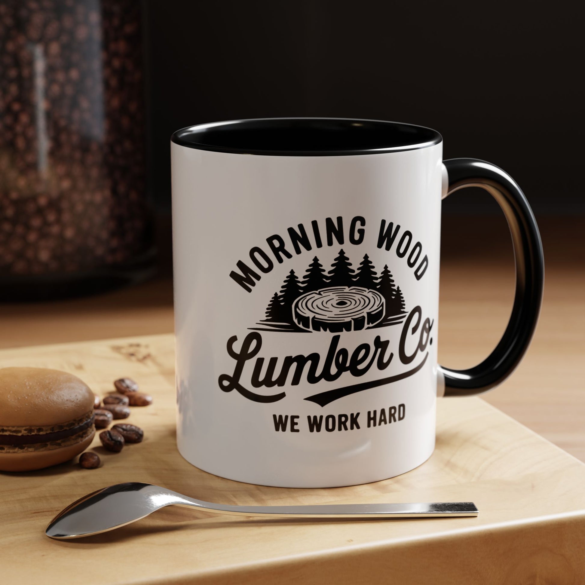 White mug with black interior and handle, featuring 'Morning Wood Lumber Co. We Work Hard' text and graphic, on a wooden surface with coffee beans and a spoon.