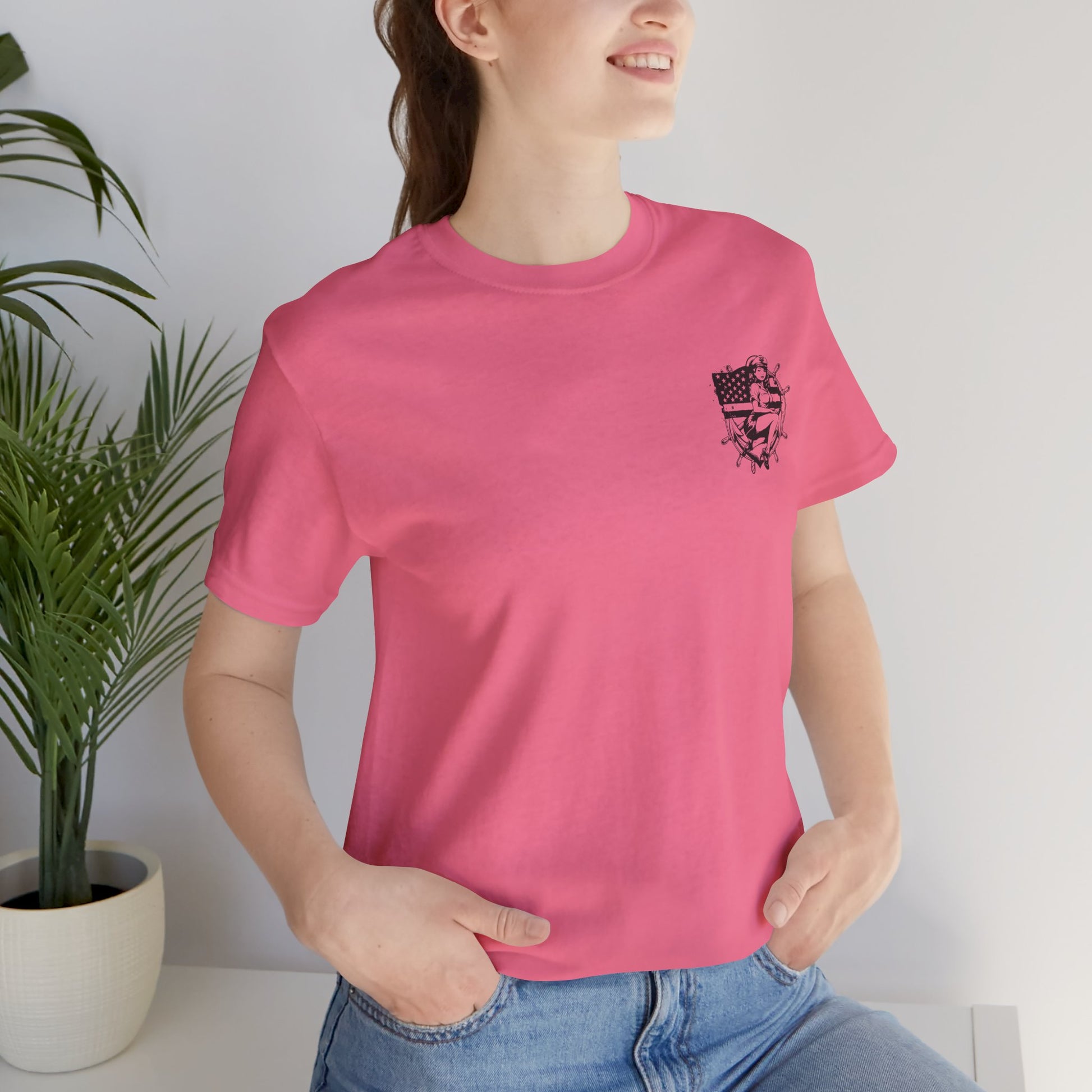 Person wearing a pink t-shirt with a graphic design, standing indoors next to a plant.