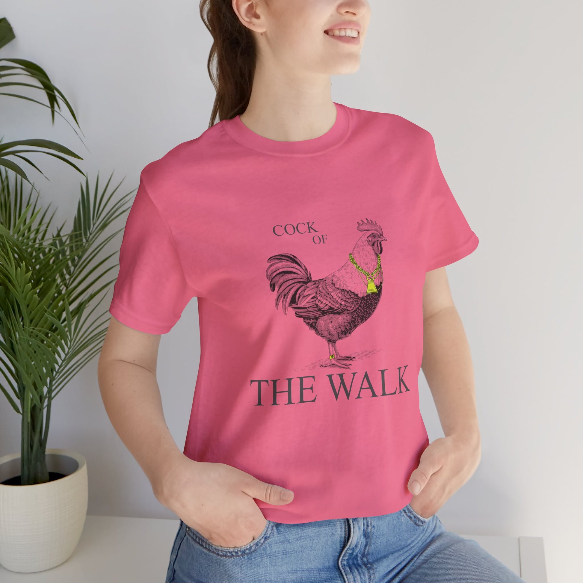 Person wearing a pink t-shirt with a rooster graphic and text, standing indoors.
