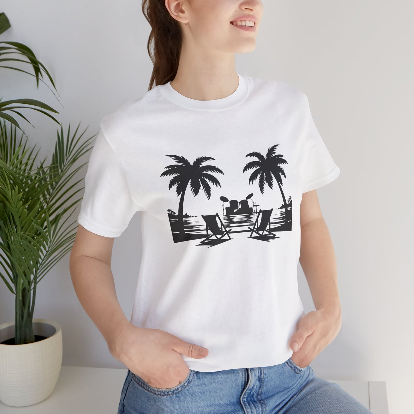 Person wearing a white t-shirt with black palm tree and beach chair design, standing indoors next to a plant.