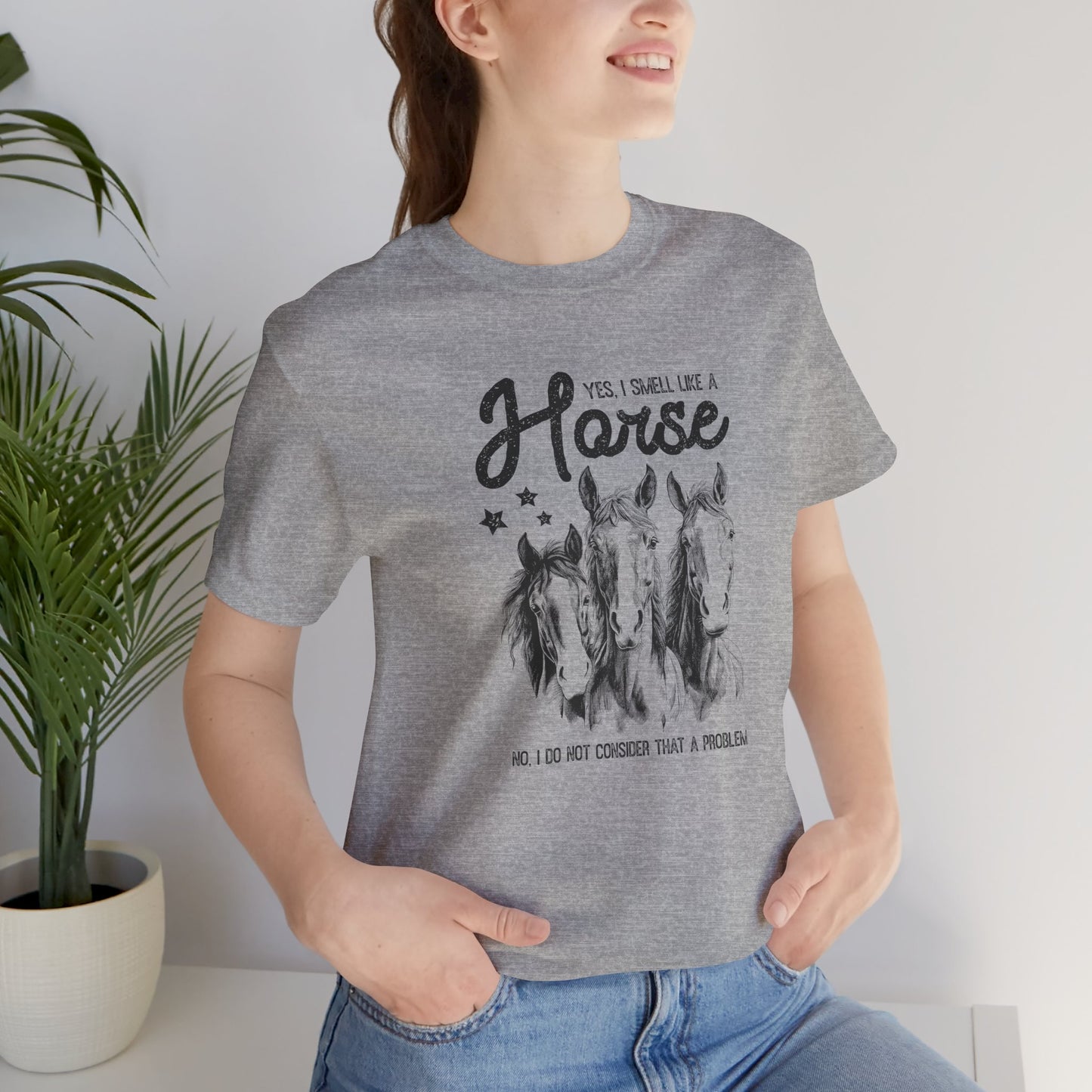 Person wearing a gray t-shirt with horse graphic and text, standing indoors next to a plant.