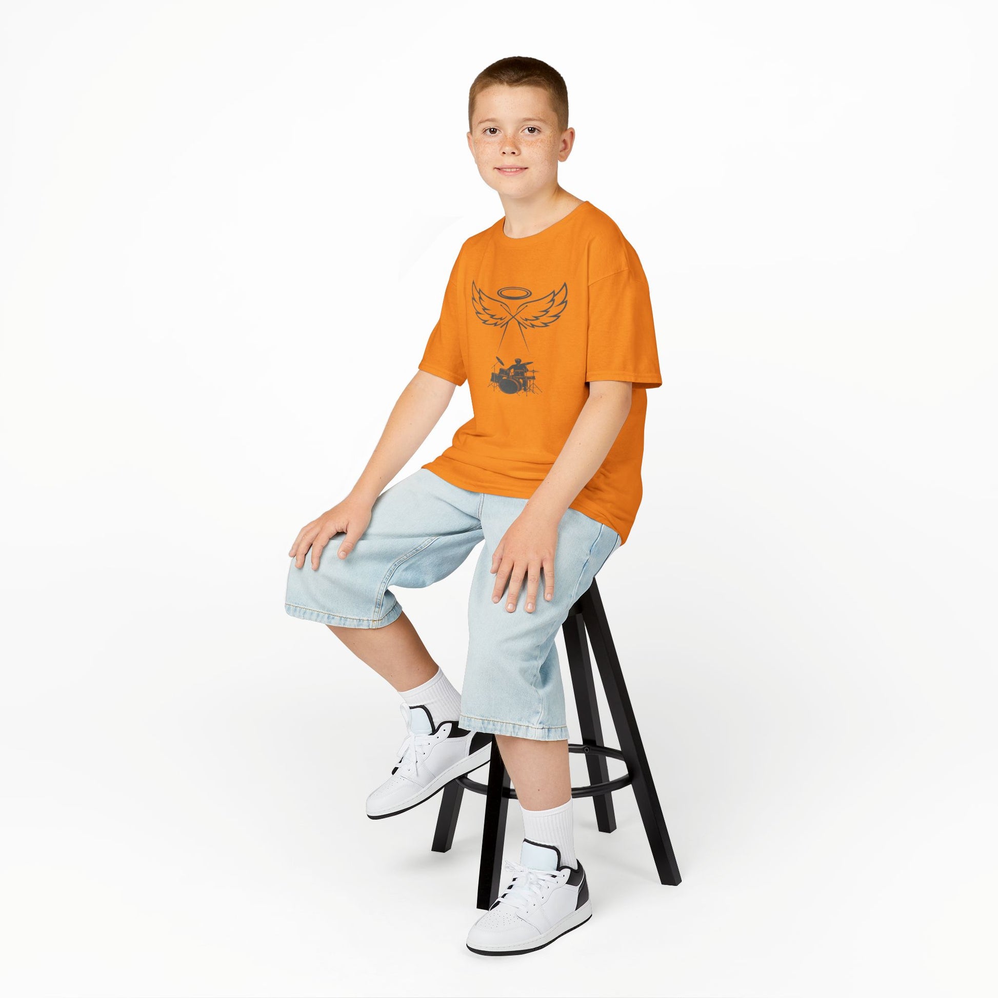 Child sitting on a stool wearing an orange t-shirt and light blue shorts on a white background