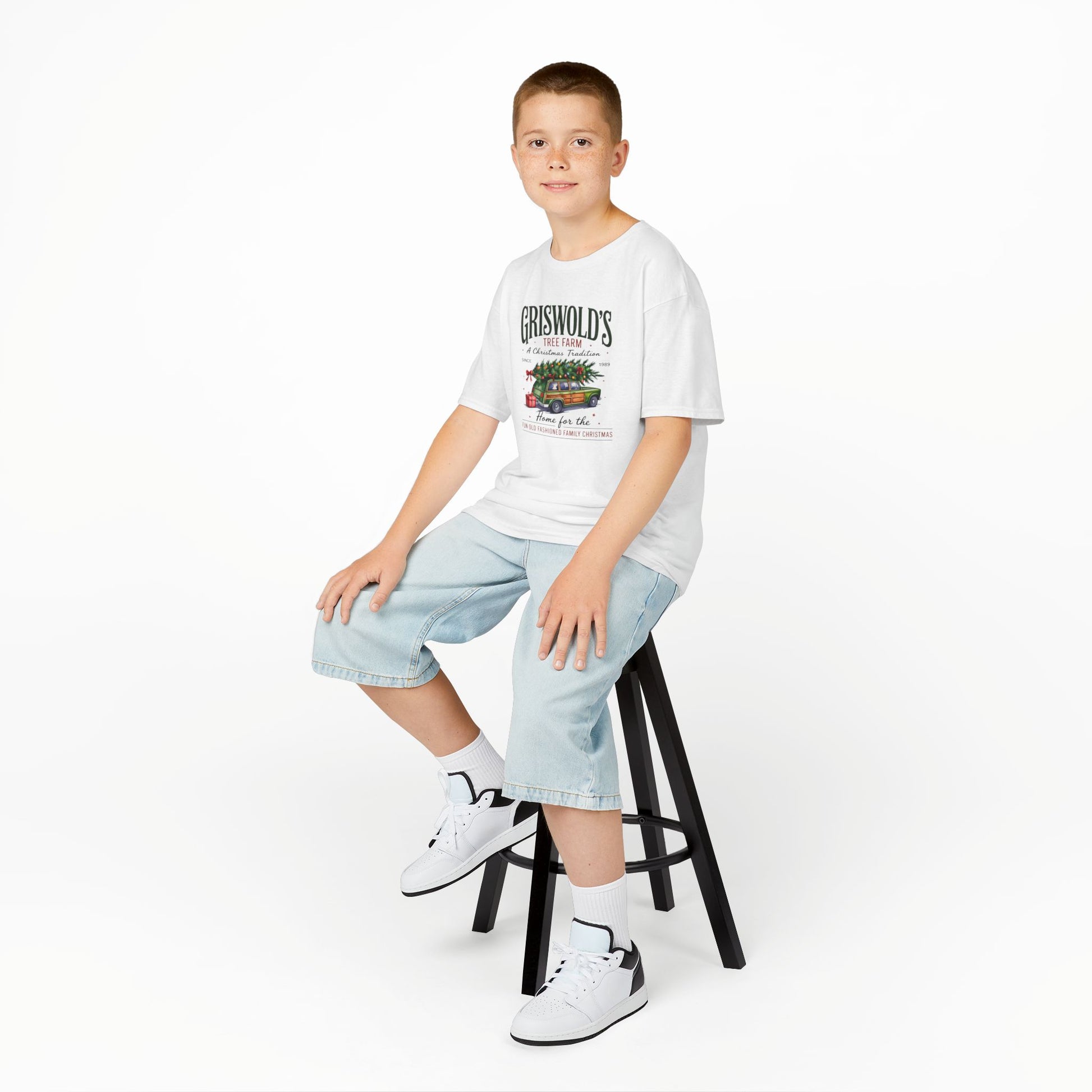 Young boy sitting on a stool wearing a white t-shirt with text and graphics, light blue jeans, and white sneakers.