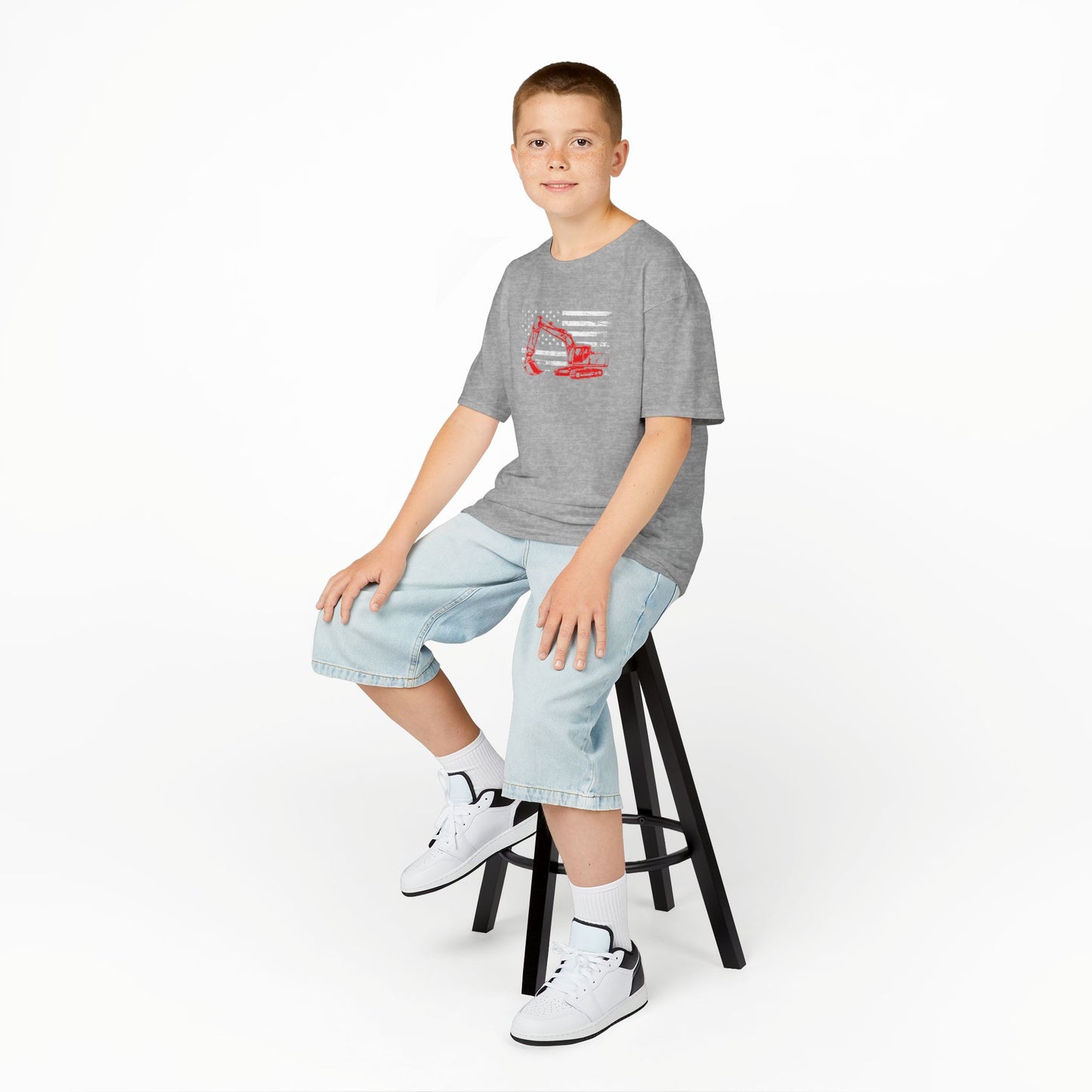 Child sitting on a stool wearing a gray t-shirt with a logo and light blue shorts on a white background