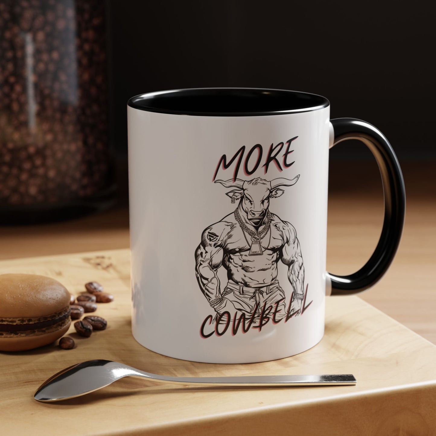 Mug with a graphic of a muscular bull and text 'More Cowbell' on a wooden surface with coffee beans and a spoon.