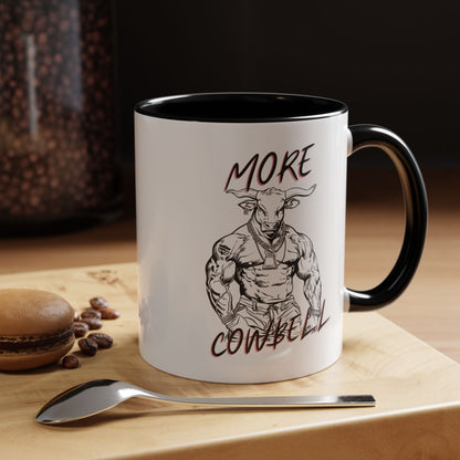 Mug with a graphic of a muscular bull and text 'More Cowbell' on a wooden surface with coffee beans and a spoon.