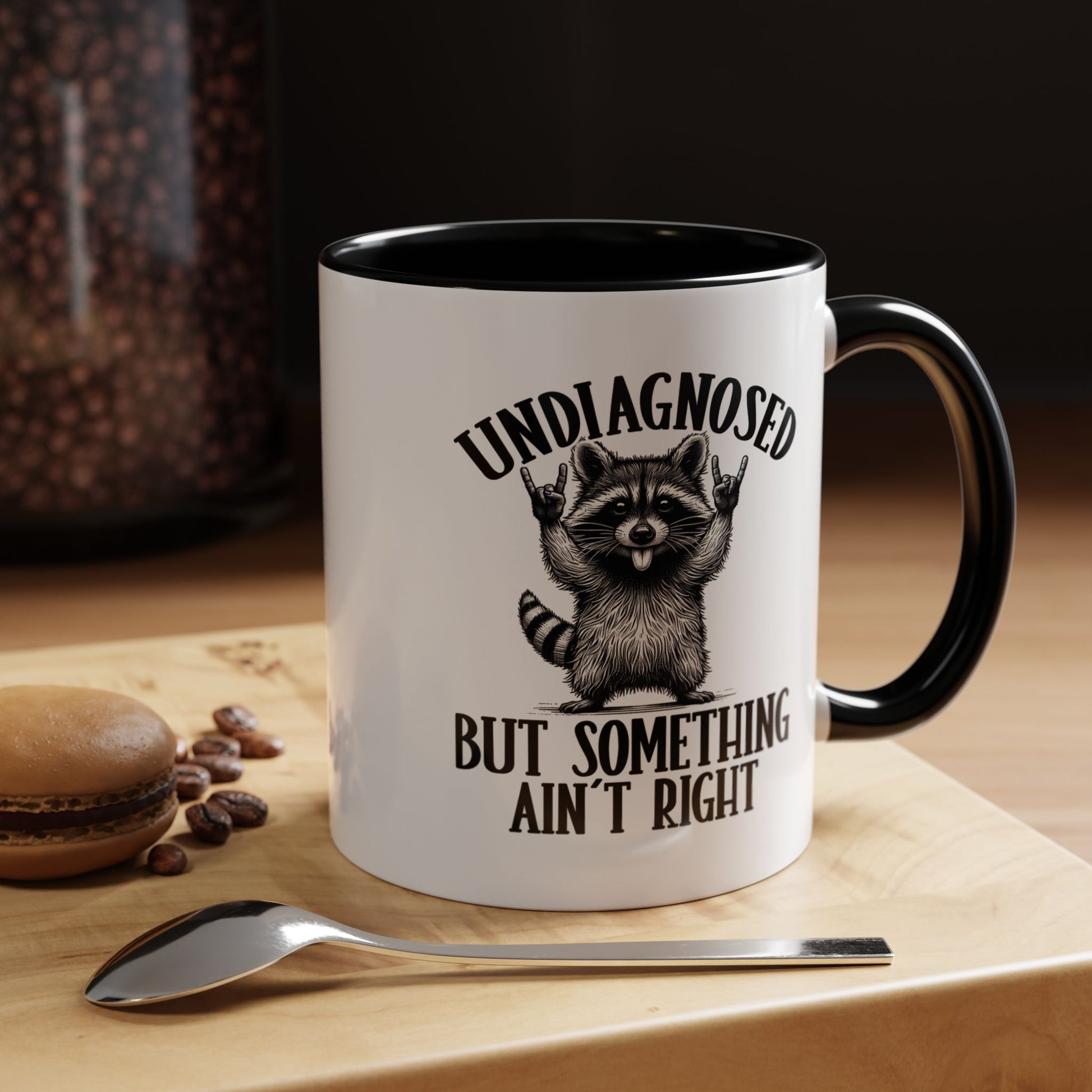 White mug with black interior and handle, featuring a raccoon graphic and text, on a wooden surface with coffee beans and a spoon.