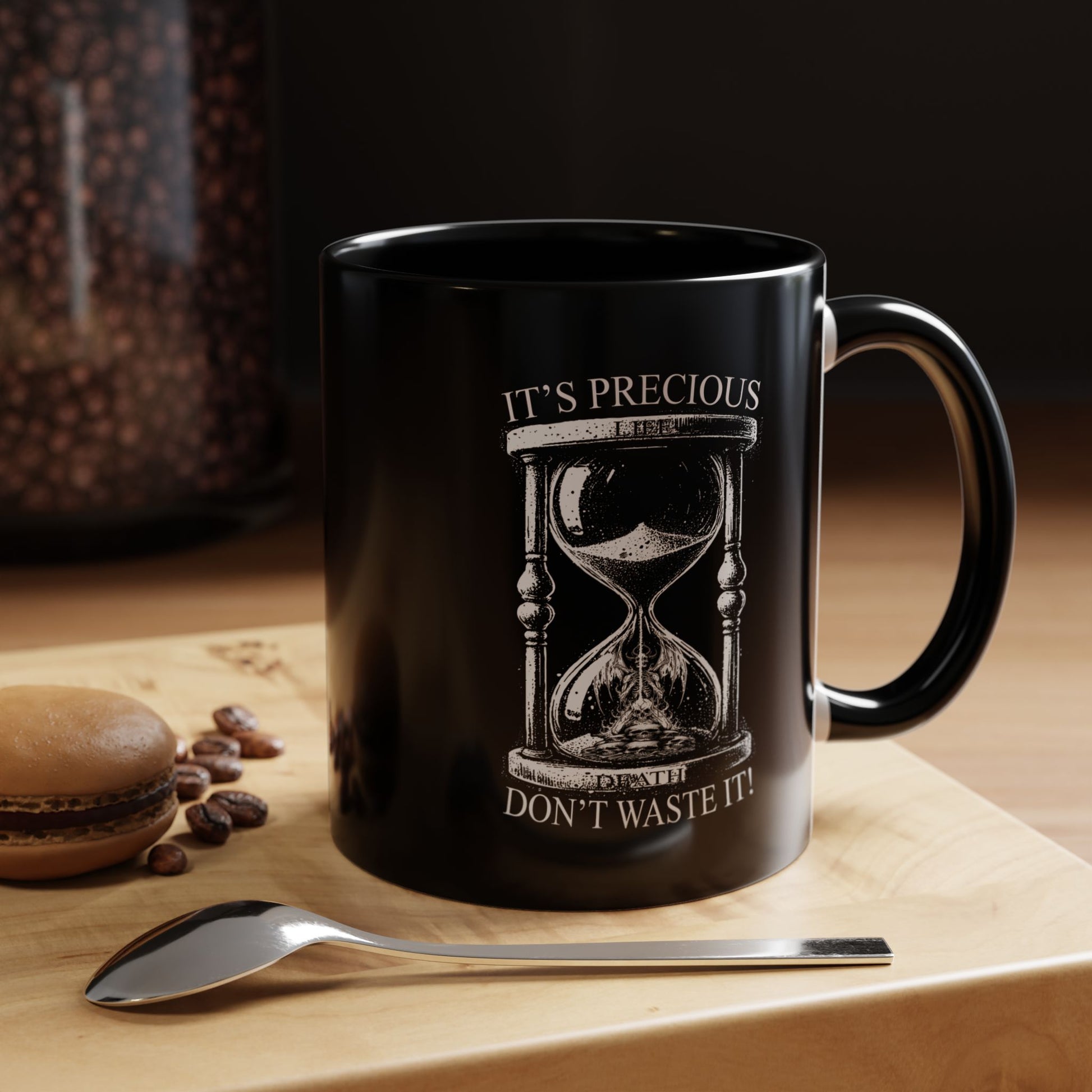 Black mug with hourglass design and text on a wooden surface with coffee beans and a spoon.