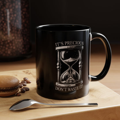Black mug with hourglass design and text on a wooden surface with coffee beans and a spoon.