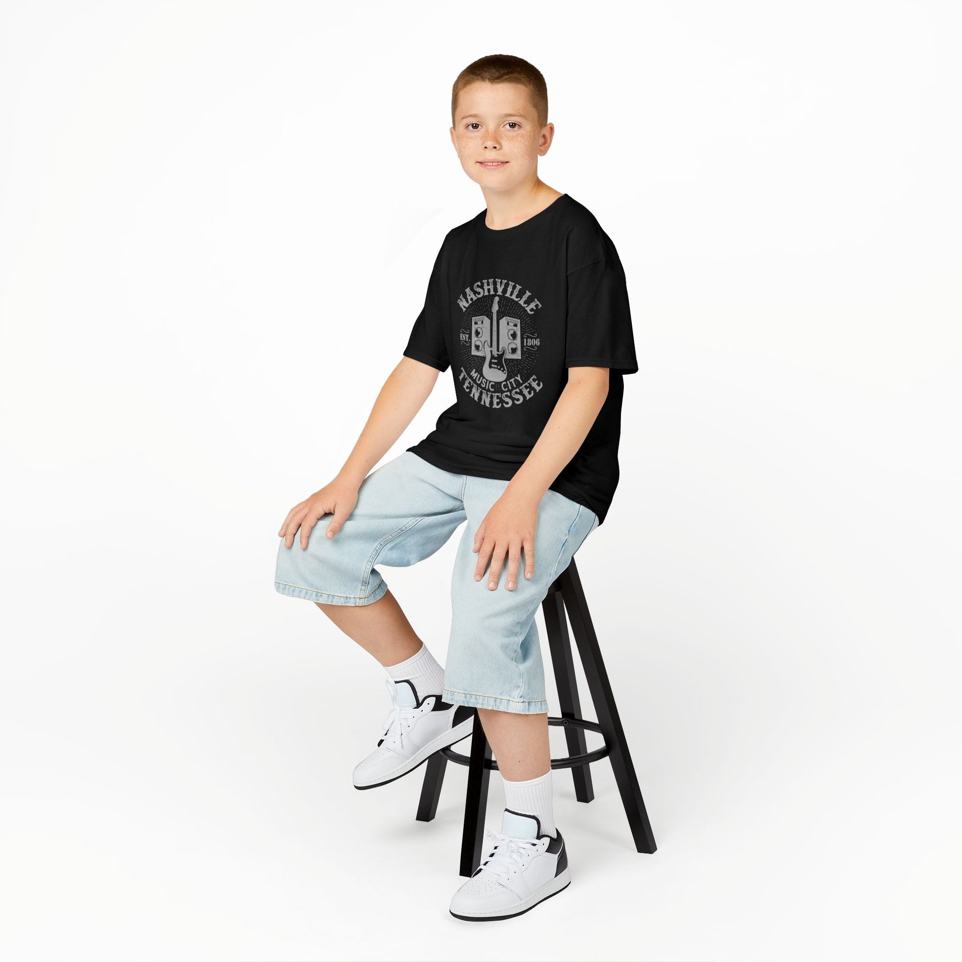 Young person sitting on a stool wearing a black t-shirt with a graphic design, light blue jeans, and white sneakers.