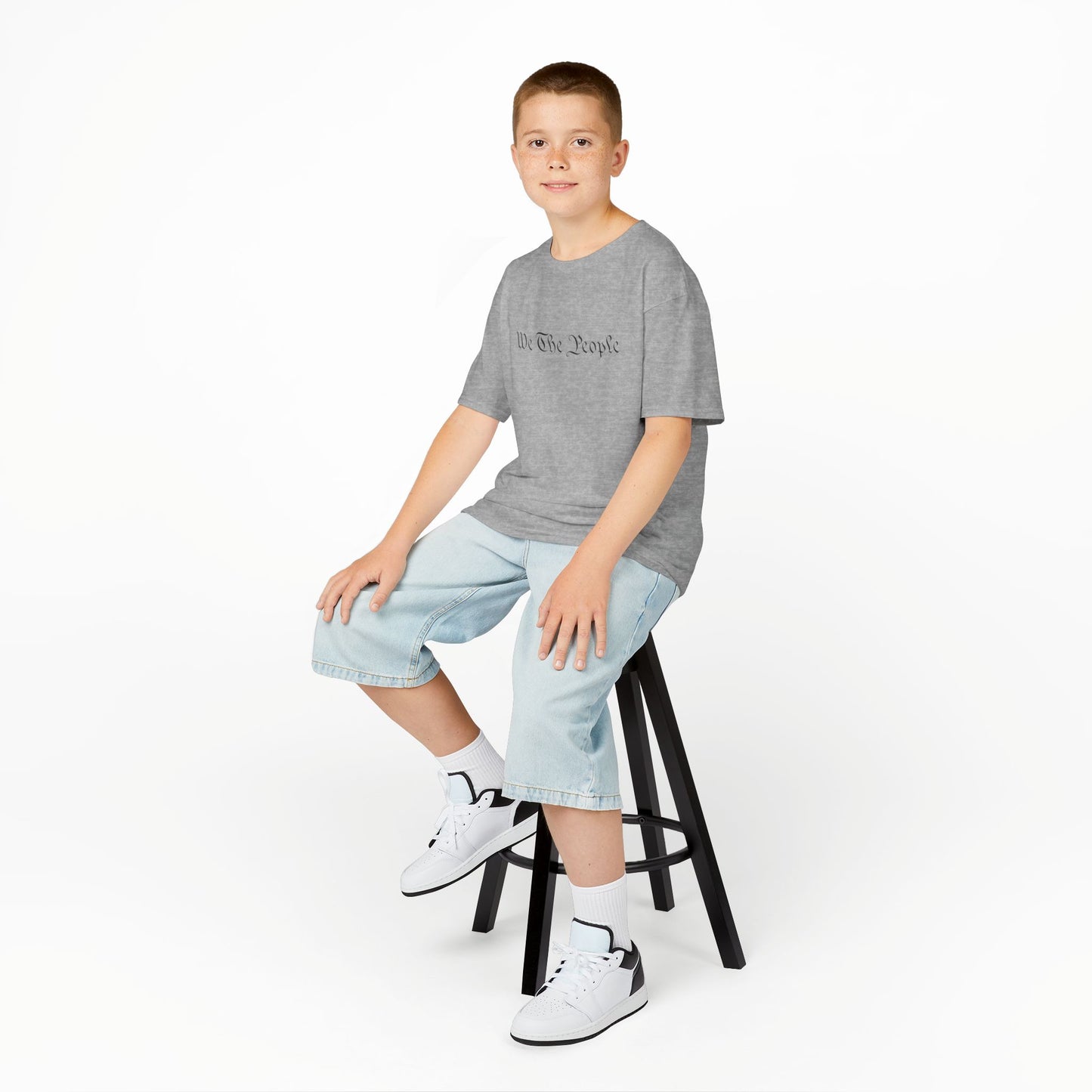 Young boy sitting on a stool wearing a gray t-shirt, light blue jeans, and white sneakers on a white background